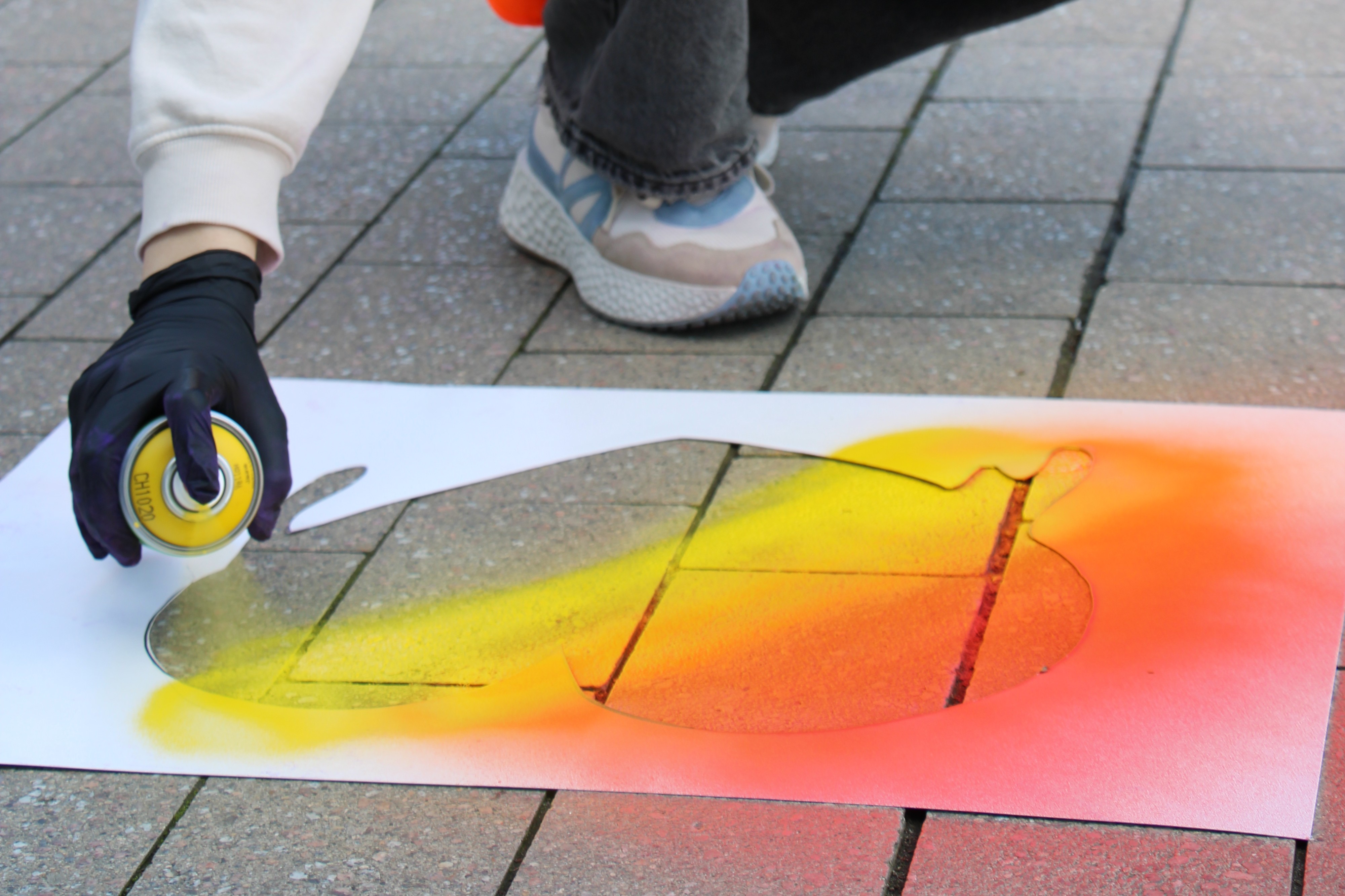 Eine hockende Person, von der Füße und Hände zu sehen sind, sprüht ein buntes Herz auf Straßenpflaster.
